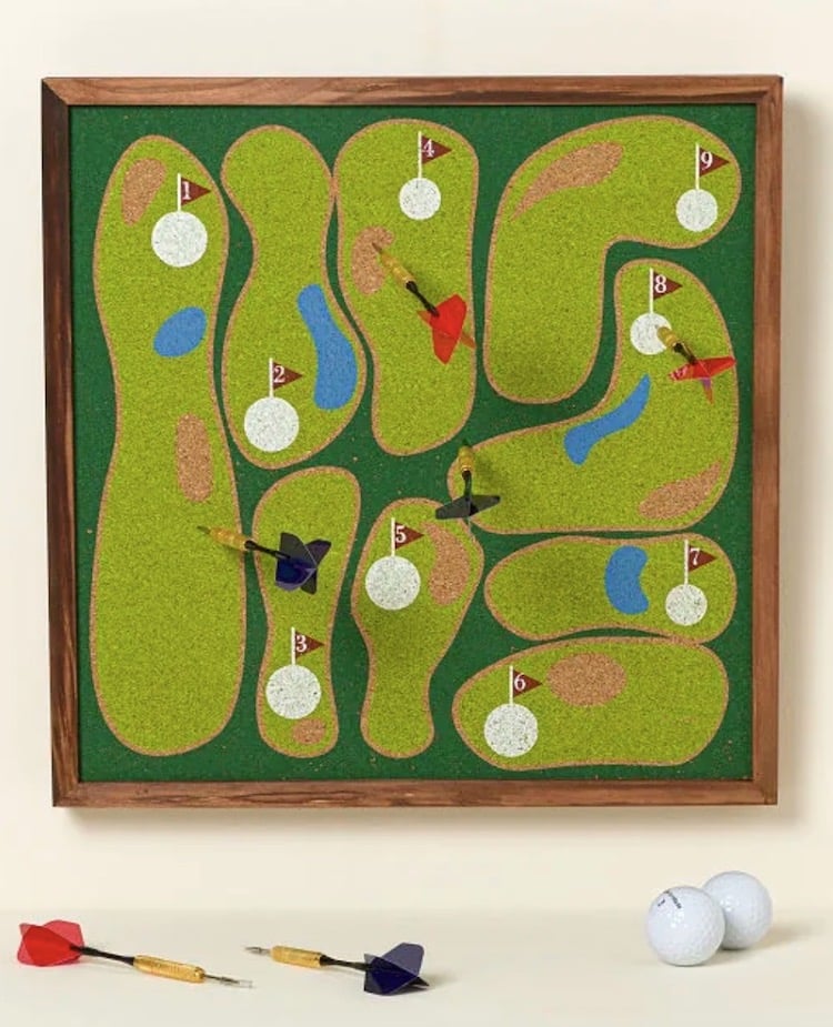 A framed corkboard shaped like a mini golf course with numbered holes, flags, and golf balls makes a fun gift idea for those who are hard to shop for. Colored darts decorate the board, with extra darts and golf balls resting on the surface below.