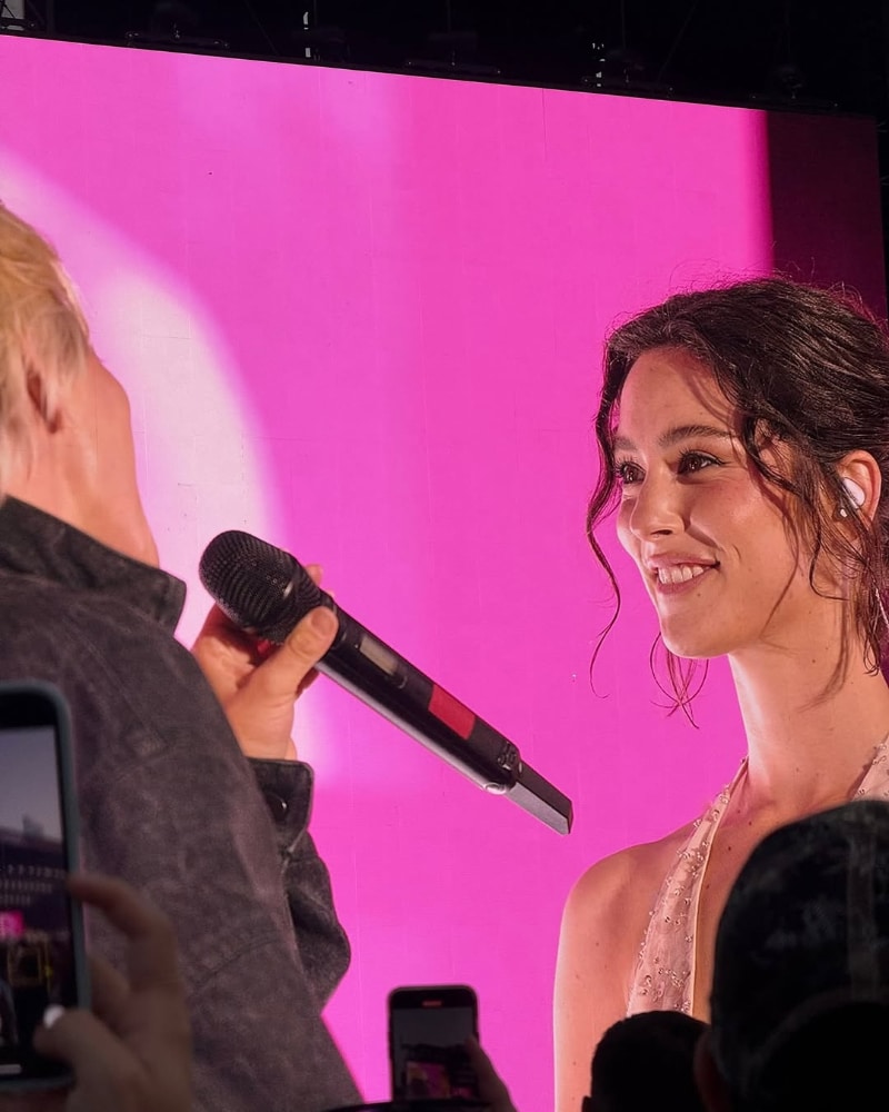 Two people are on stage in front of a large bright pink screen. One holds a microphone, singing or speaking to the other, who is smiling with her hair styled in loose curls and wearing a sparkly sleeveless top.