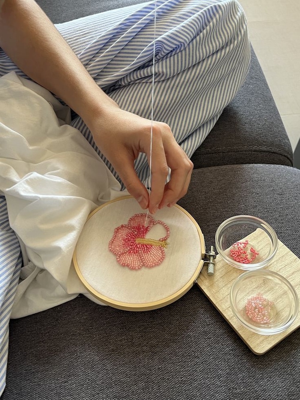 A person in striped pajamas is embroidering a pink flower on white fabric using a wooden hoop—a perfect example of grandmacore hobbies. Small glass bowls with pink and green beads rest nearby on a wooden board.