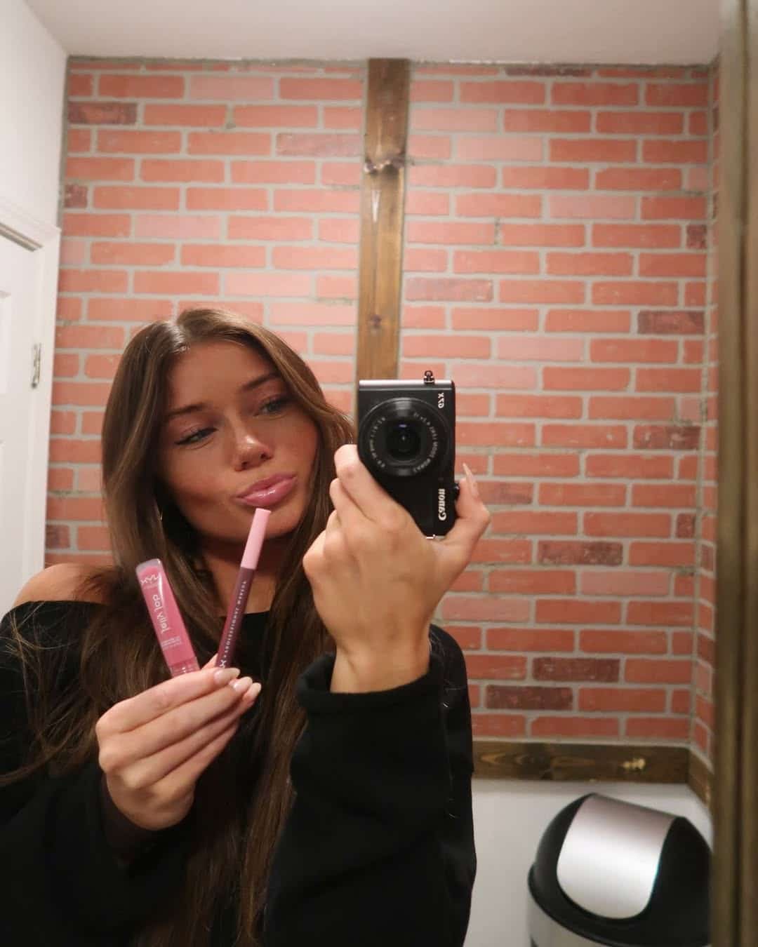 A woman with long brown hair takes a mirror selfie in a bathroom with brick walls, holding two pink lip products and pouting her lips—showcasing makeup tips. A silver trash can is visible in the corner.