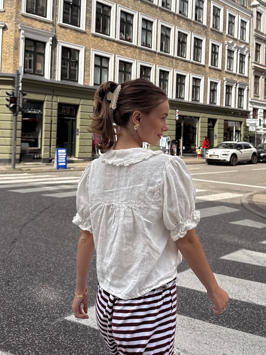 A woman with a claw clip hair accessory wears a white blouse and striped pants, walking across a city street at a crosswalk. The background shows historic buildings, cars, and a traffic light—perfect inspiration for everyday styling tips.