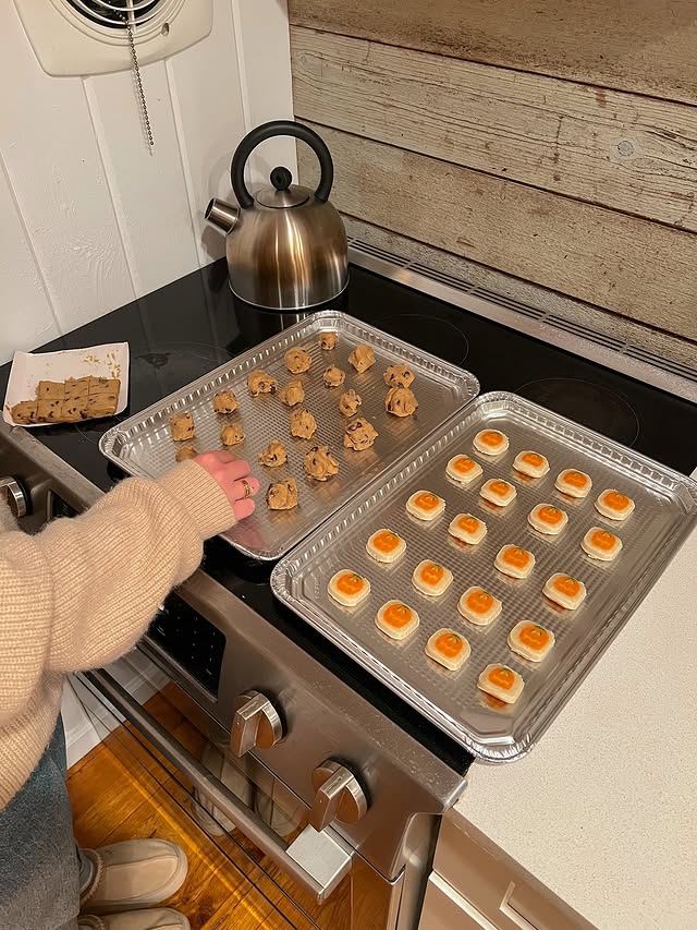 A person prepares Sweet Fall Treats, placing cookie dough on a baking tray in a cozy kitchen. Two trays with raw cookies rest on the stove, and a metal kettle sits on the back burner. The person wears a beige sweater and blue jeans.