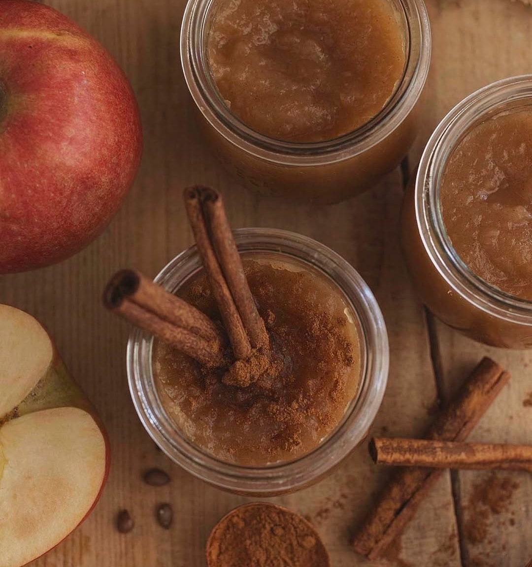 Three jars of applesauce topped with cinnamon sticks and ground cinnamon sit on a wooden surface, surrounded by a whole apple, a sliced apple, and loose cinnamon—perfect for Hanukkah recipes or festive holiday gatherings.
