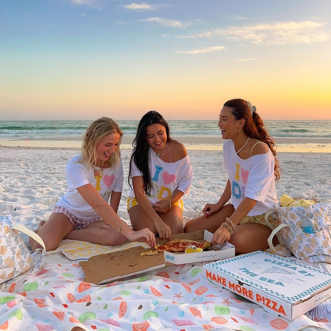 Three women sit on a beach at sunset, laughing and sharing pizza. Wearing matching I ♥ DAD shirts, they swap relationship advice as they relax on colorful blankets with pizza boxes, the ocean stretching out behind them.