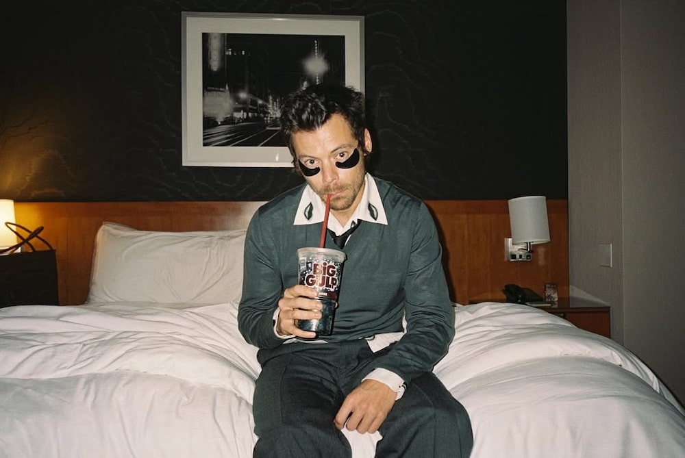 A man with dark hair sits on a bed in a hotel room, wearing a green sweater, white collared shirt, and a black eye mask. He holds a large drink cup and looks at the camera. A framed photo hangs on the wall behind him.