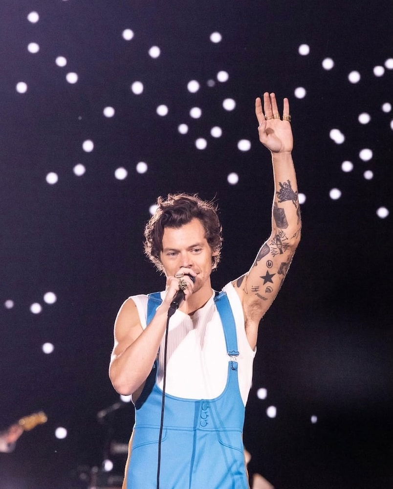 A singer in a blue jumpsuit holds a microphone and raises one arm, performing on stage with bright white lights glowing in the dark background during Harry Styles’ Together Together Tour.