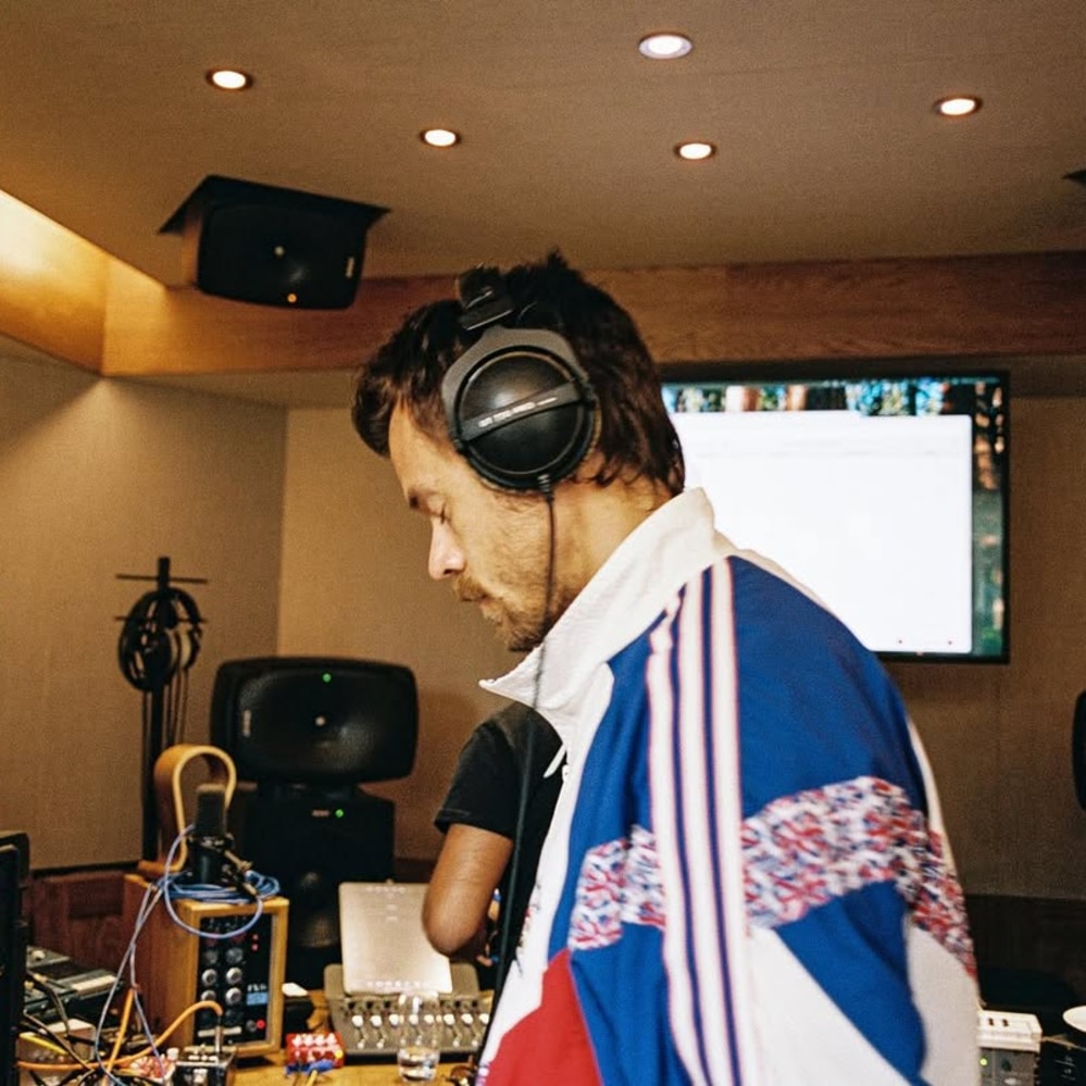A man wearing large headphones and a colorful track jacket stands in a recording studio with audio equipment, speakers, and a screen in the background.