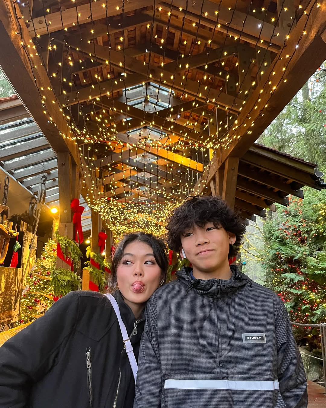 Two young people stand under a wooden pavilion adorned with festive string lights and greenery. Amid holiday decorations and trees, the girl playfully sticks out her tongue, sharing a lighthearted moment while dealing with family drama.