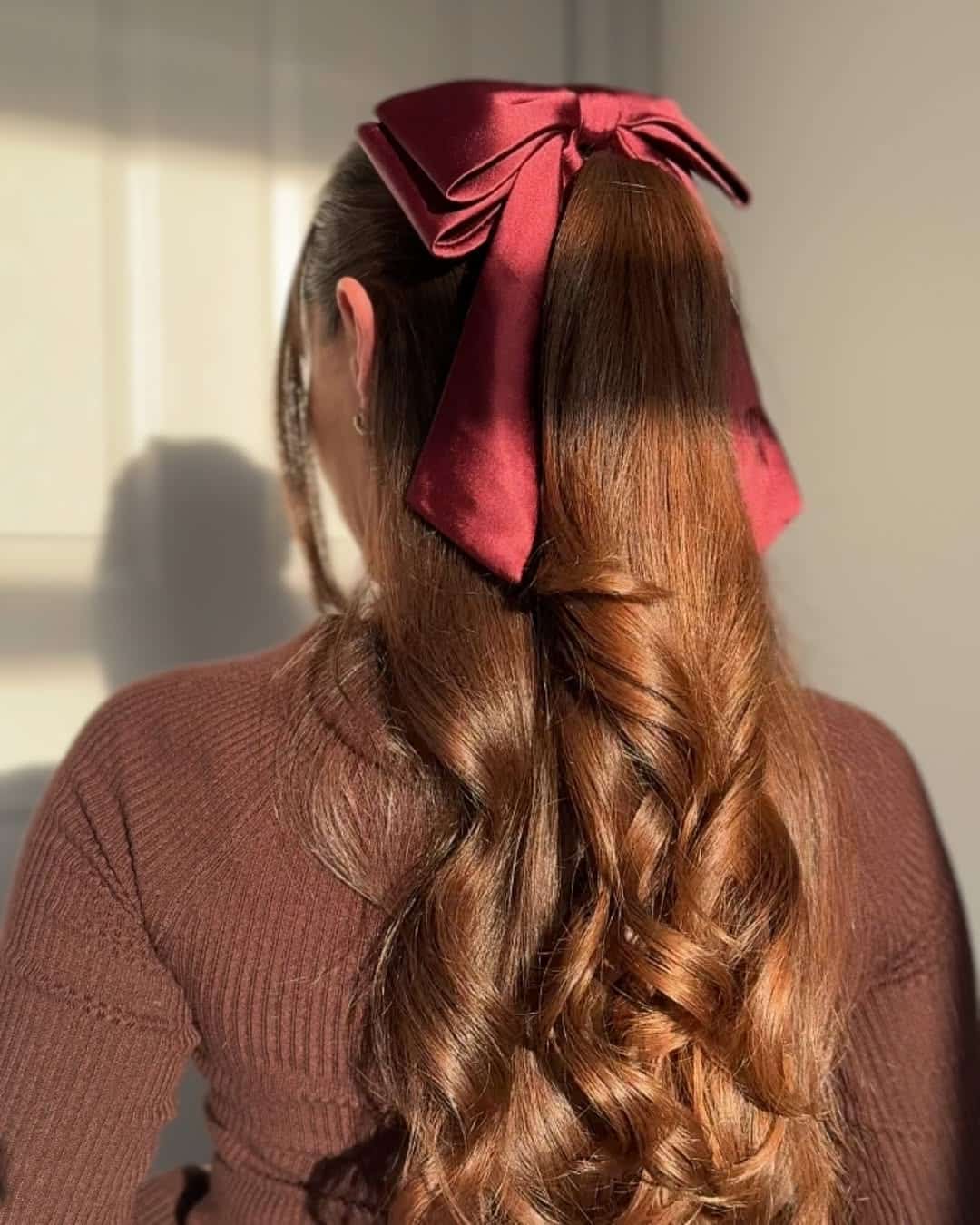 A woman with long, wavy brown hair wears a large, dark red satin bow—an elegant touch perfect for holiday hairstyles. She stands facing away from the camera in soft, natural light, dressed in a brown top.
