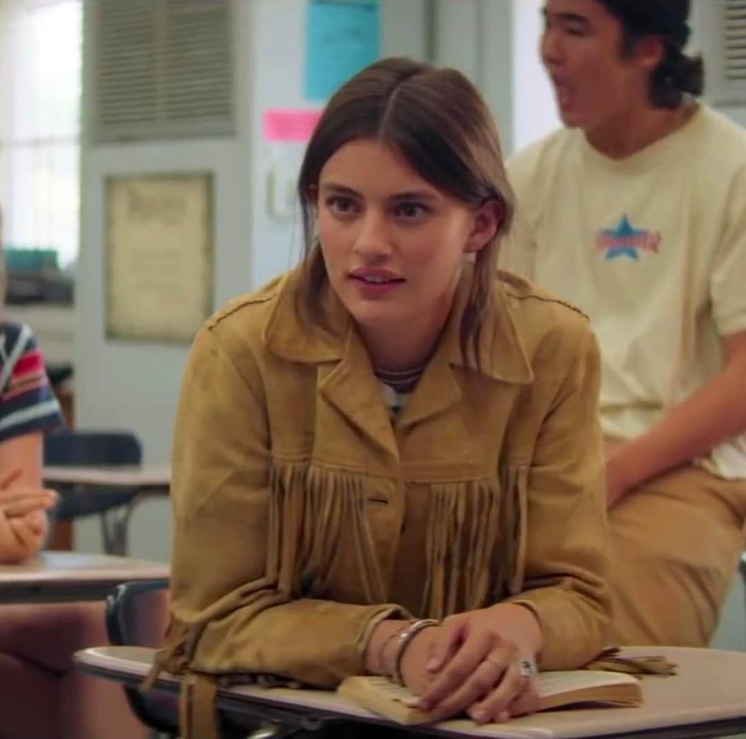 A young woman with long brown hair wearing a tan fringed jacket sits at a desk in a classroom, looking attentive, as if awaiting a Valentine's Day surprise. Other students are visible in the background.