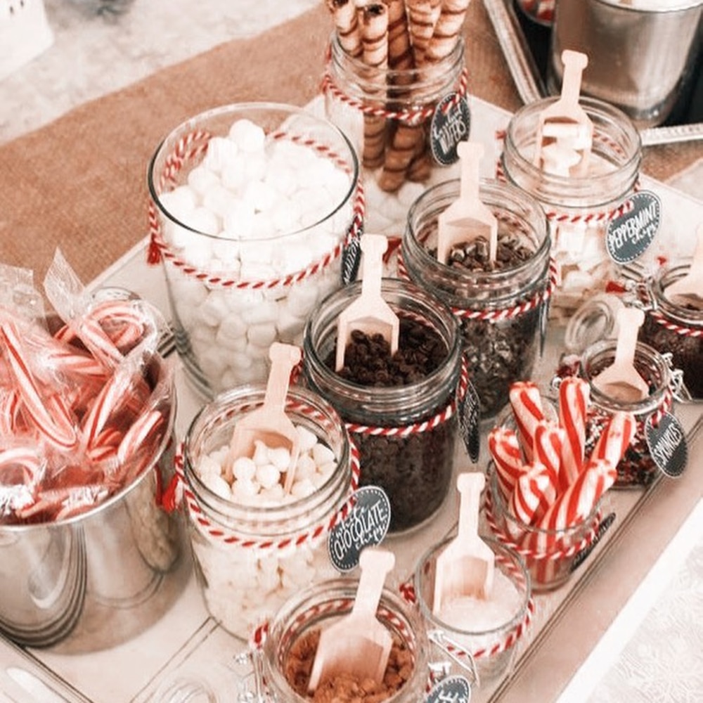 A display of jars filled with hot chocolate toppings like marshmallows, chocolate chips, and peppermint candies, each with a small wooden scoop, on a festive table.