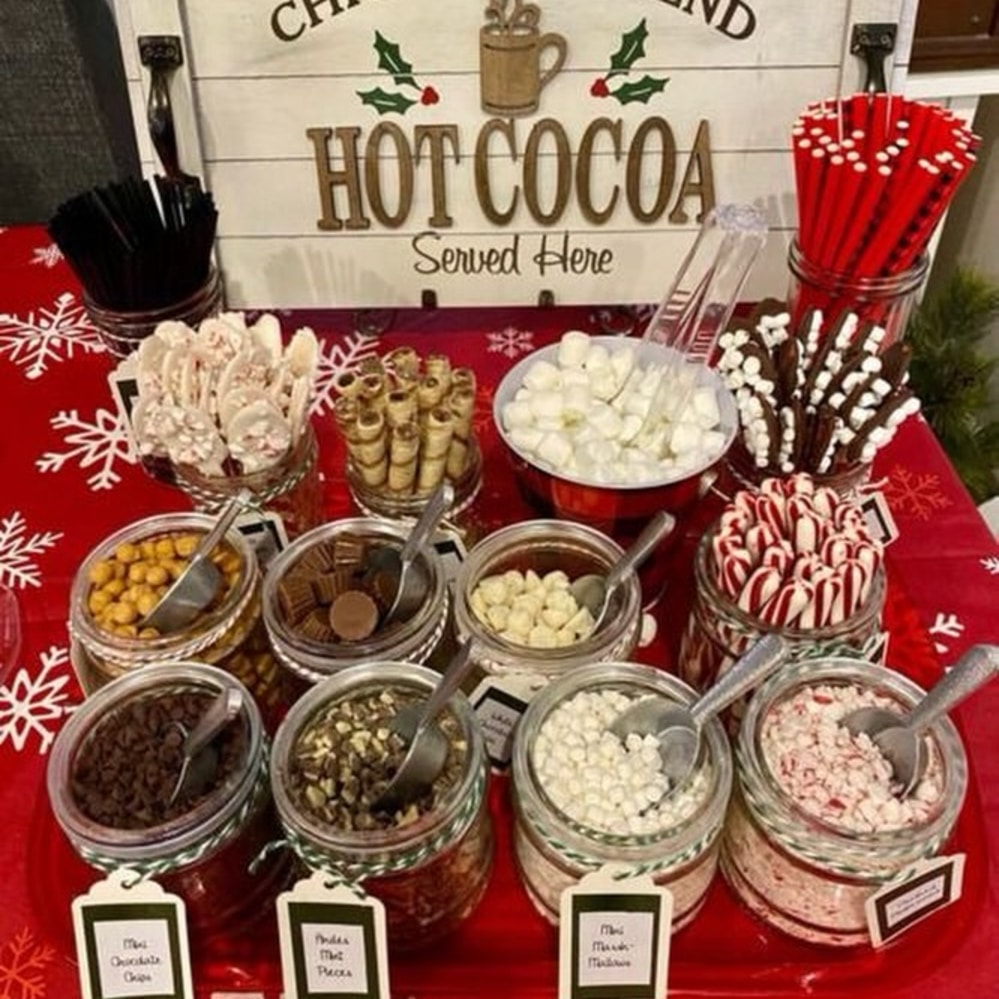 A festive hot cocoa bar with jars of toppings like marshmallows, peppermint candies, chocolate chips, wafer rolls, and chocolate pieces on a red snowflake tablecloth, with a “Hot Cocoa Served Here” sign in the background.
