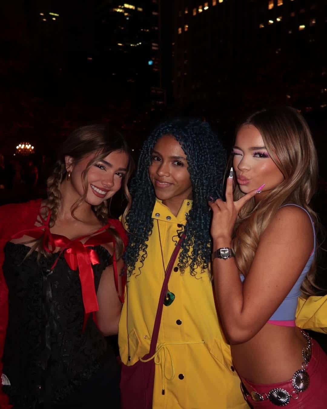 Three women in colorful costumes and stunning makeup pose and smile at night in an outdoor, city setting with tall buildings. One rocks blue braids and a yellow jacket, while another gives a peace sign—perfect for sharing beauty hacks or makeup tips!.