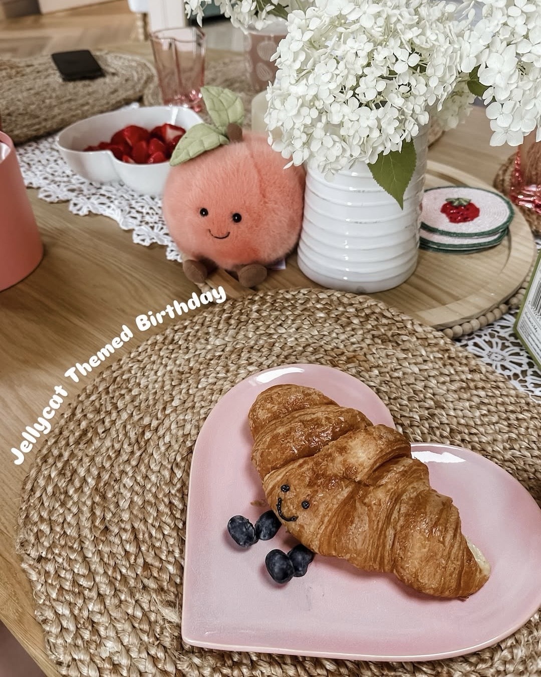 A croissant decorated with blueberries to look like a Jellycat plush sits on a pink heart-shaped plate. Nearby are white flowers, a plush peach toy, and strawberries—perfect decor to Host Jellycat Party or an Ultimate Jellycat Party. Text: Jellycat Themed Birthday.