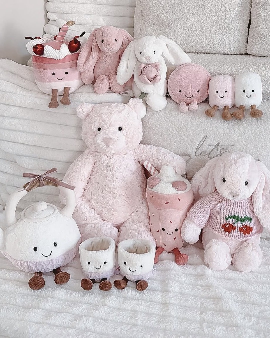 A group of pastel pink and white plush toys, including bunnies, a teddy bear, a teapot, milk cartons, and desserts with smiling faces, are arranged on a white bed—ready to host a cozy Jellycat party.