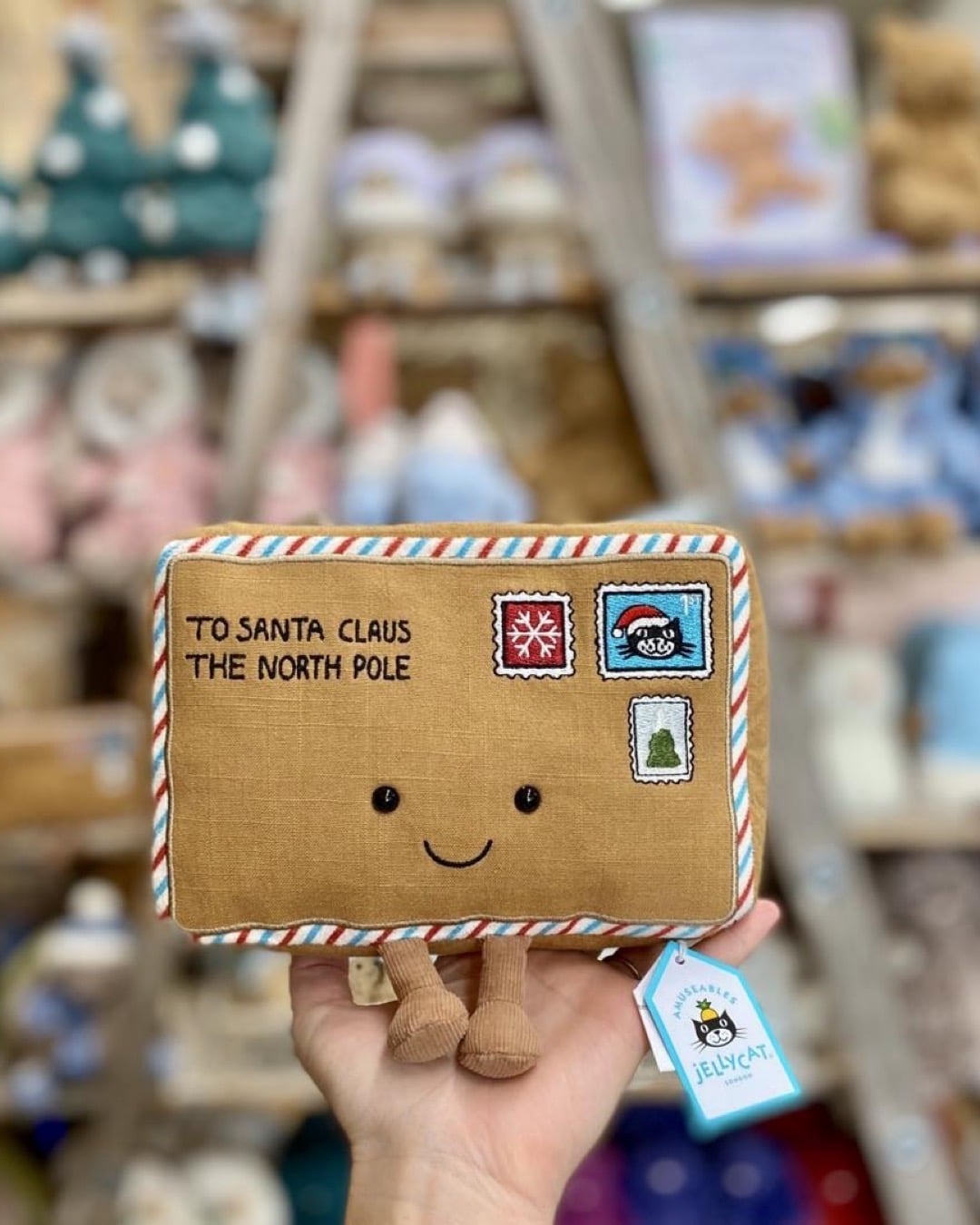 A hand holds a plush toy shaped like a letter, with embroidered stamps and text reading To Santa Claus The North Pole. Perfect for your next Jellycat party, the letter has a smiling face, legs, and blurred holiday decorations in the background.