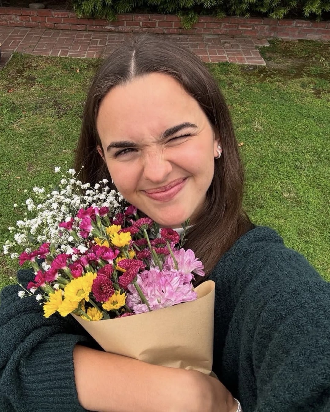 A young woman with long brown hair smiles and winks while holding a bouquet of colorful flowers wrapped in brown paper, standing on a green lawn with a brick path and bushes in the background.