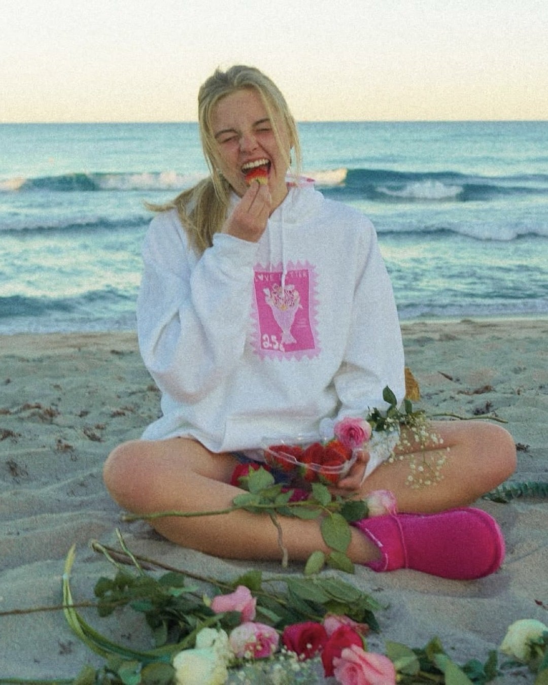 A young woman sits on the beach wearing a white hoodie and pink slippers, surrounded by scattered flowers. Smiling with her eyes closed while eating a strawberry, she embodies the Single Girl’s Guide to Surviving Valentine’s Day by the ocean.
