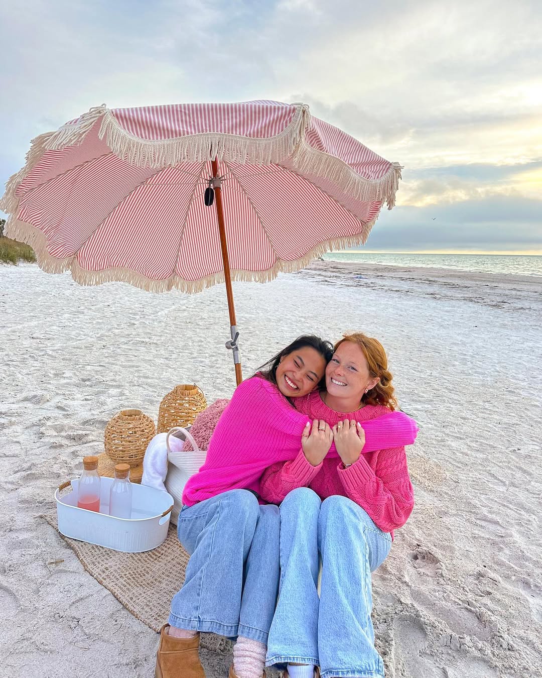 Two women in bright pink sweaters and jeans hug and smile under a pink striped beach umbrella on a sandy shore—a perfect Single Girl’s Guide moment. A picnic basket and drinks sit on a mat beside them. The sky is partly cloudy.