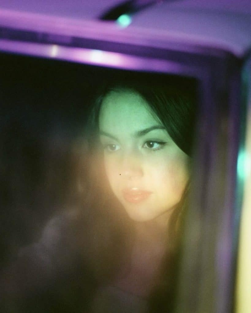 A young woman with dark hair looks thoughtfully through a glass window, illuminated by soft green and purple lights that create a dreamy, atmospheric effect.
