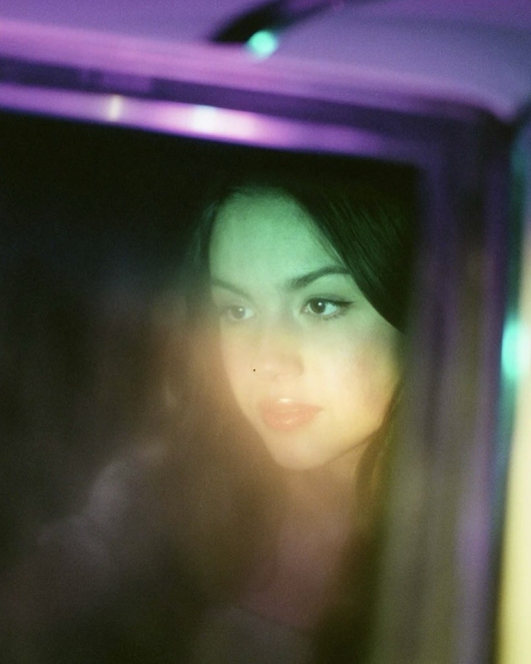 A young woman with dark hair looks thoughtfully through a glass window, illuminated by soft green and purple lights that create a dreamy, atmospheric effect.