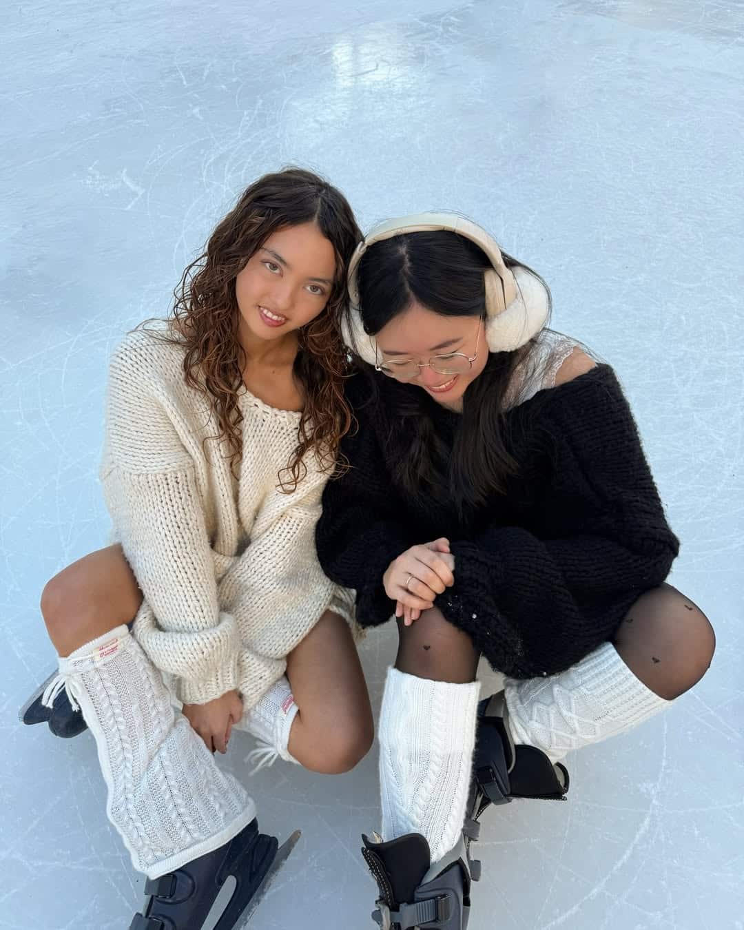 Two young women in cozy sweaters and leg warmers sit on an ice rink, smiling. With their ice skates and cheerful poses, they enjoy winter activities together—capturing a joyful moment of family bonding.