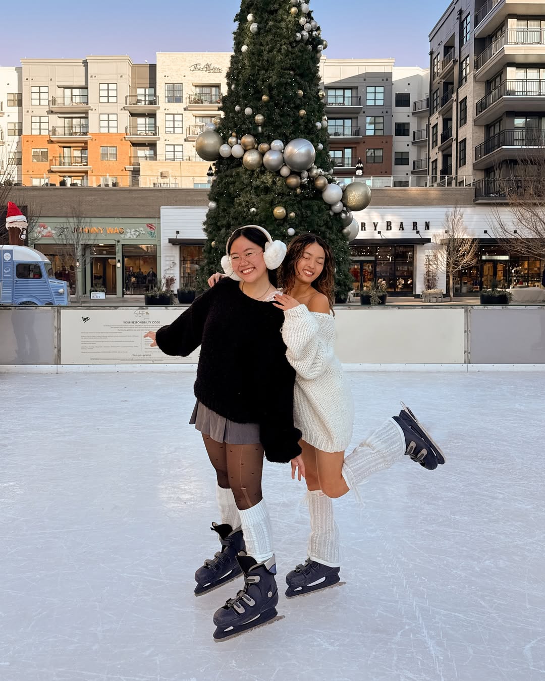 Two women in winter outfits smile and pose on an outdoor ice skating rink, enjoying winter activities together in front of a large decorated Christmas tree, with buildings and shops behind them. One woman lifts her leg playfully.