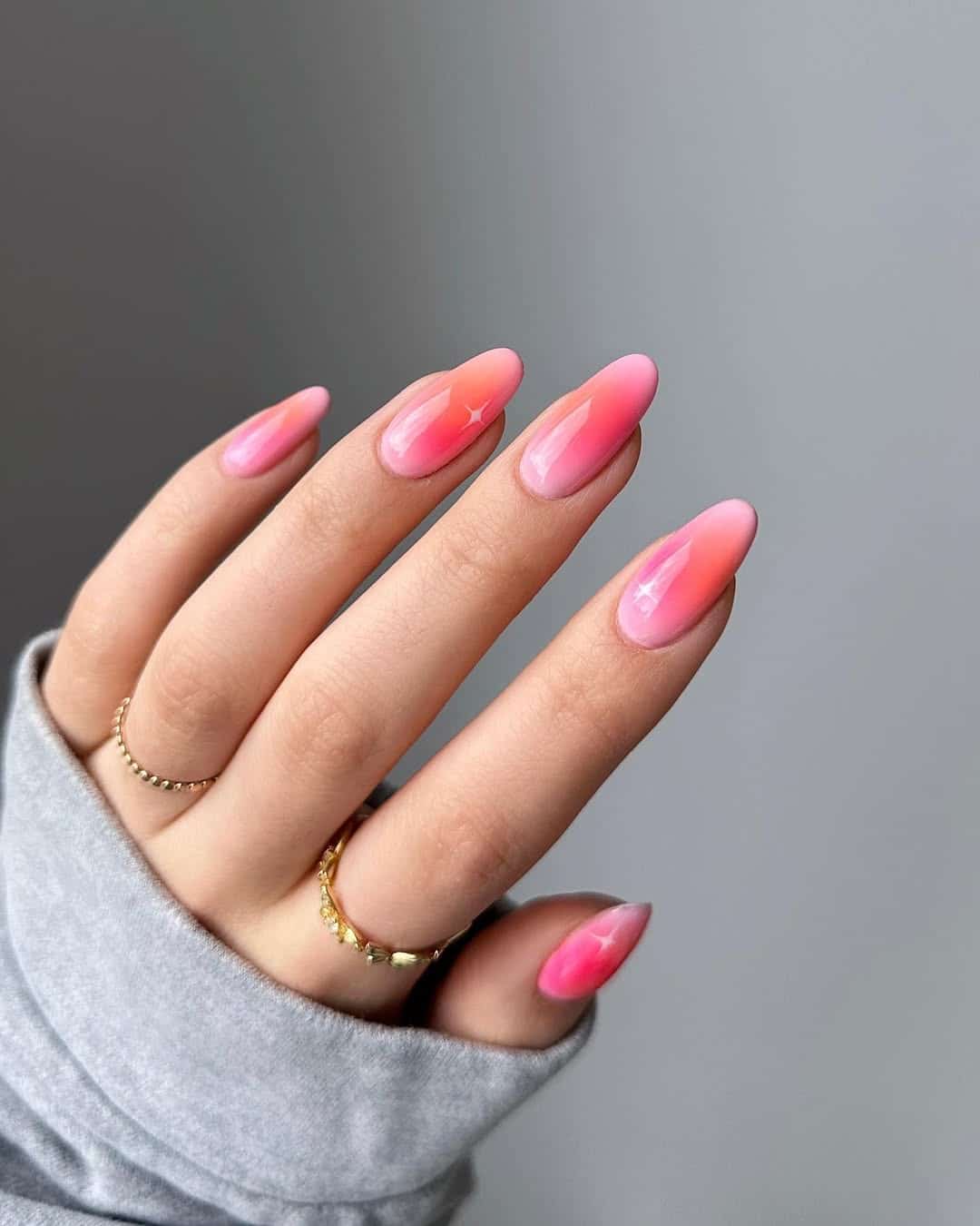 A hand with long, almond-shaped nails painted in a pink ombre design, featuring a glossy finish and subtle star accents. The person is wearing gold rings and a light gray sleeve.