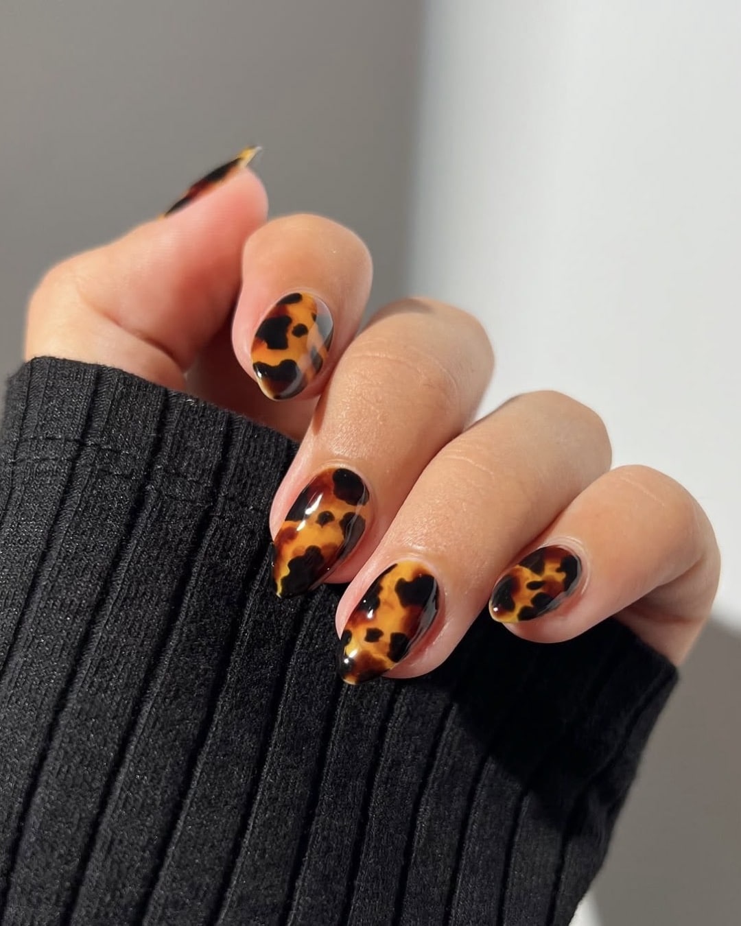 A hand with almond-shaped nails painted in a glossy tortoiseshell pattern, holding the fingers slightly curled. The person is wearing a long-sleeved black ribbed top.
