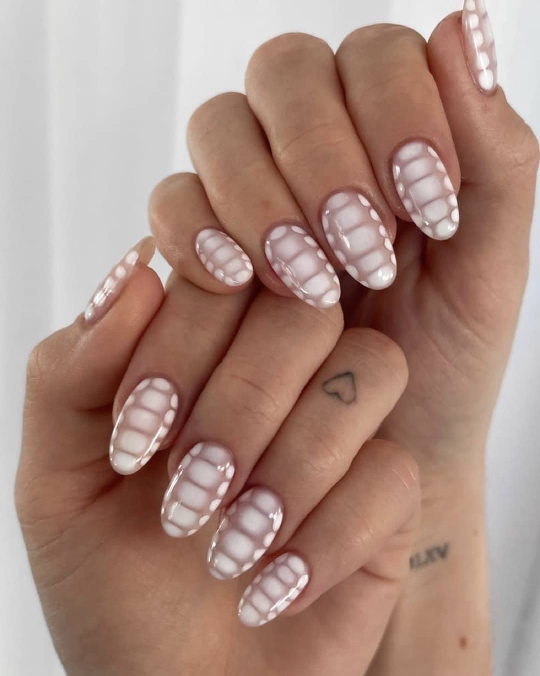 A hand with long, almond-shaped nails featuring white designs resembling teeth or a skeletal pattern. The nails are glossy, and there is a small heart tattoo on the persons finger.