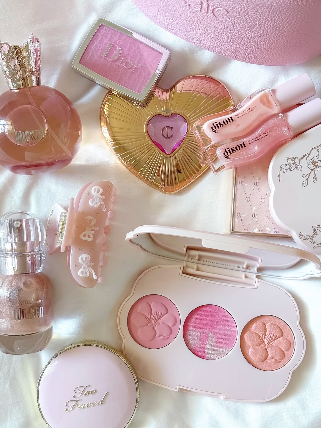 A collection of pink and gold makeup items—including blush, lip gloss, perfume bottles, a hair clip, and a compact—arranged neatly on white fabric, perfect for discovering the best beauty dupes in the beauty world.