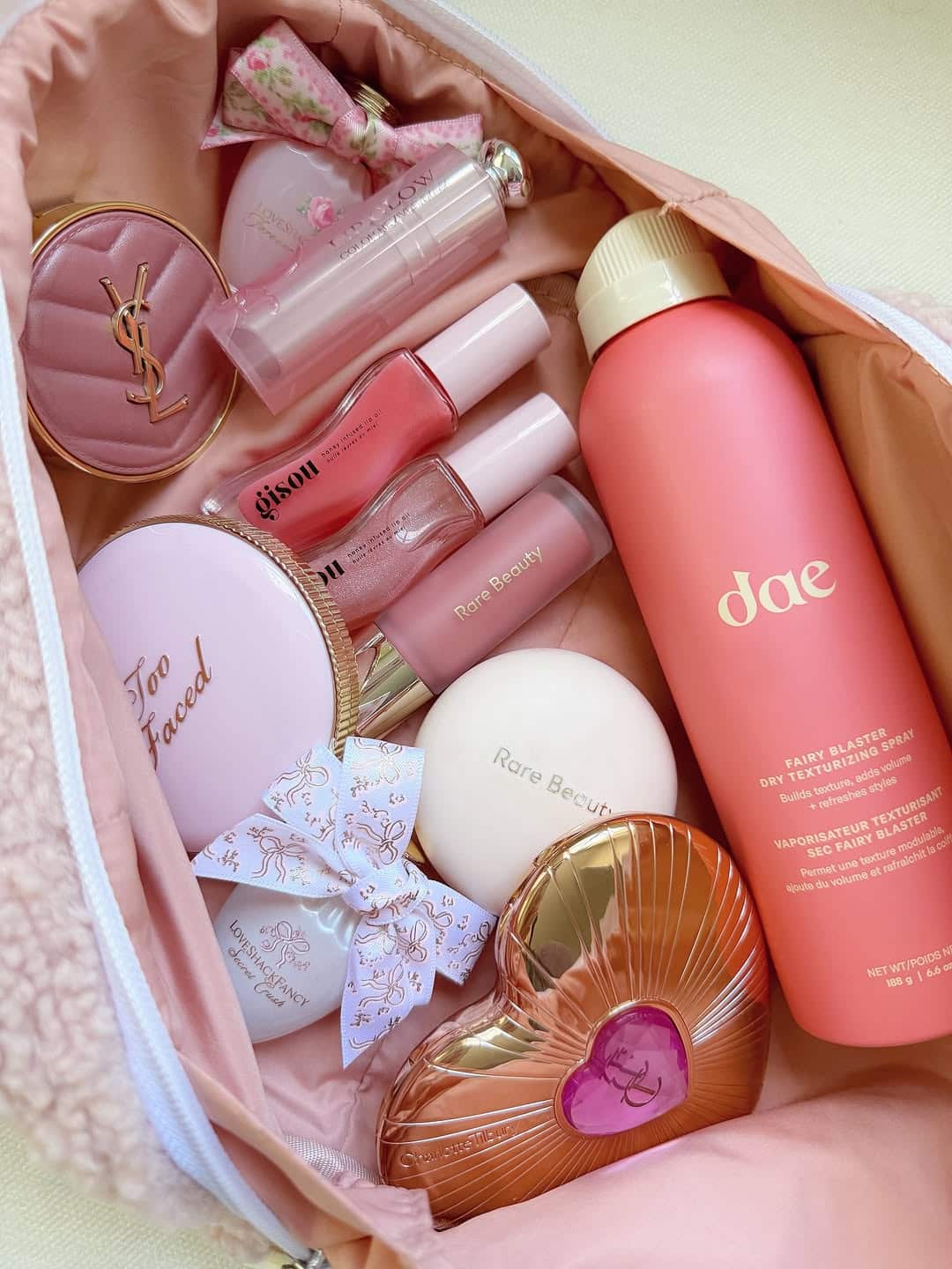 A pink makeup bag filled with beauty world favorites, including Too Faced and Rare Beauty compacts, dae dry shampoo, perfume, pink lip glosses, ribbon bows, and a heart-shaped Yves Saint Laurent item—all arranged neatly.