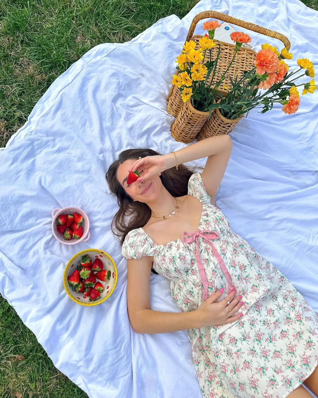 A woman in a floral dress lies on a white blanket outdoors, holding a strawberry over one eye. Beside her are bowls of strawberries, a wicker basket, and orange-yellow flowers. Grass surrounds the picnic scene.