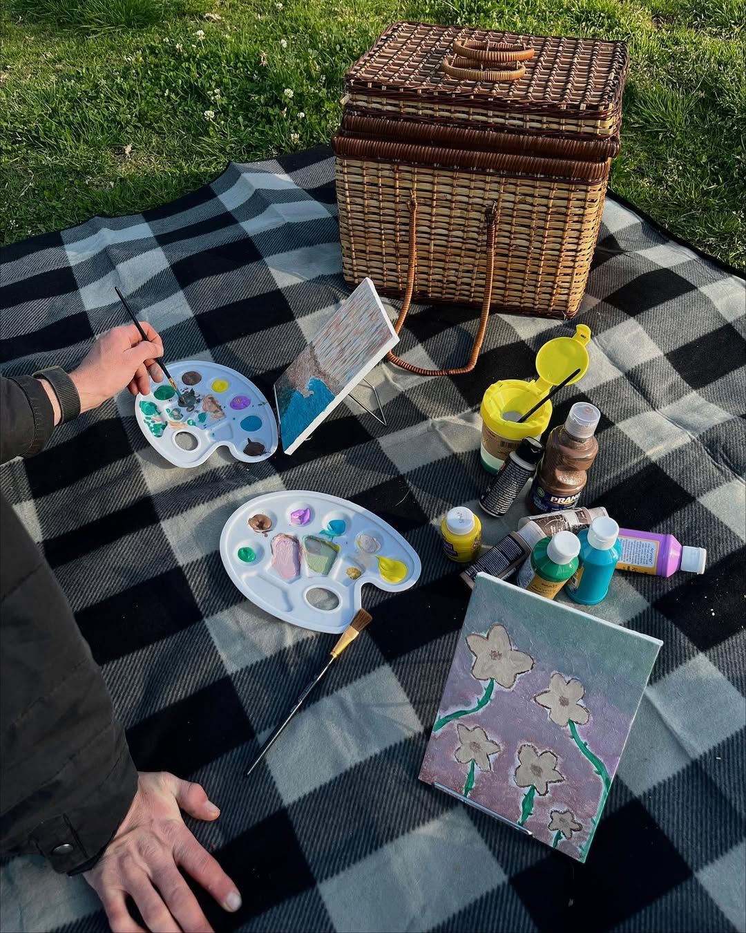 Two people painting outdoors on a checkered blanket with palettes, paint tubes, brushes, and flower artwork. A wicker picnic basket is nearby on the grass under sunlight.