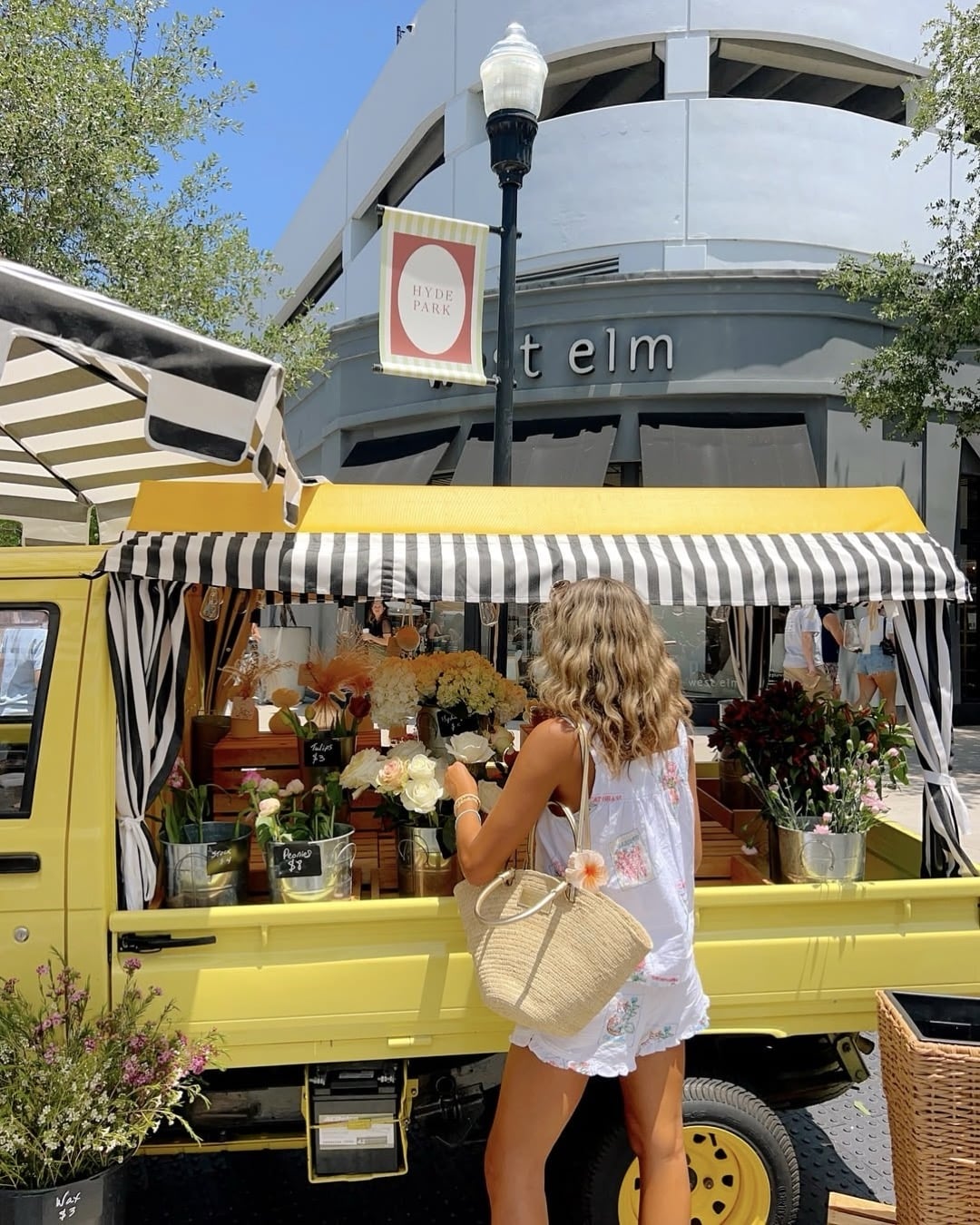 A woman in a white dress stands at a yellow flower truck with a black-and-white striped awning, selecting flowers on a sunny day, with a West Elm store and trees in the background.