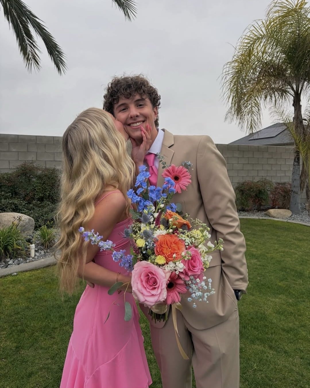 A young couple poses outside; the woman in a pink dress kisses the man, who wears a tan suit and pink tie. He smiles, holding a colorful bouquet of flowers. Palm trees and a garden are in the background.