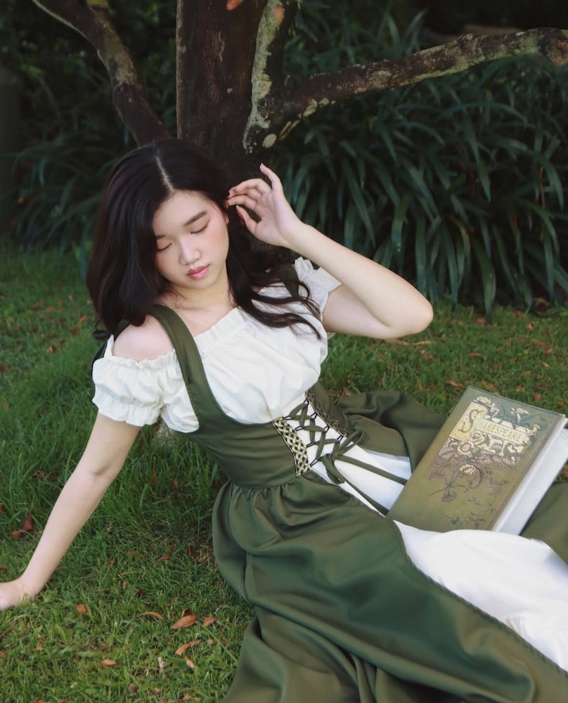 A young woman in a green and white dress sits on grass under a tree, eyes closed, with one hand to her hair. A closed, ornate book rests on her lap. Lush greenery and tree branches surround her.