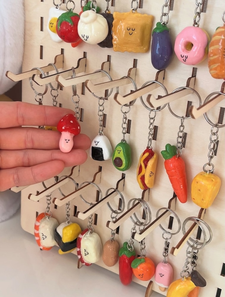 A hand holds a cute mushroom keychain among many colorful, smiling food-themed keychains, including sushi, avocado, carrot, and hot dog, displayed on a wooden stand.
