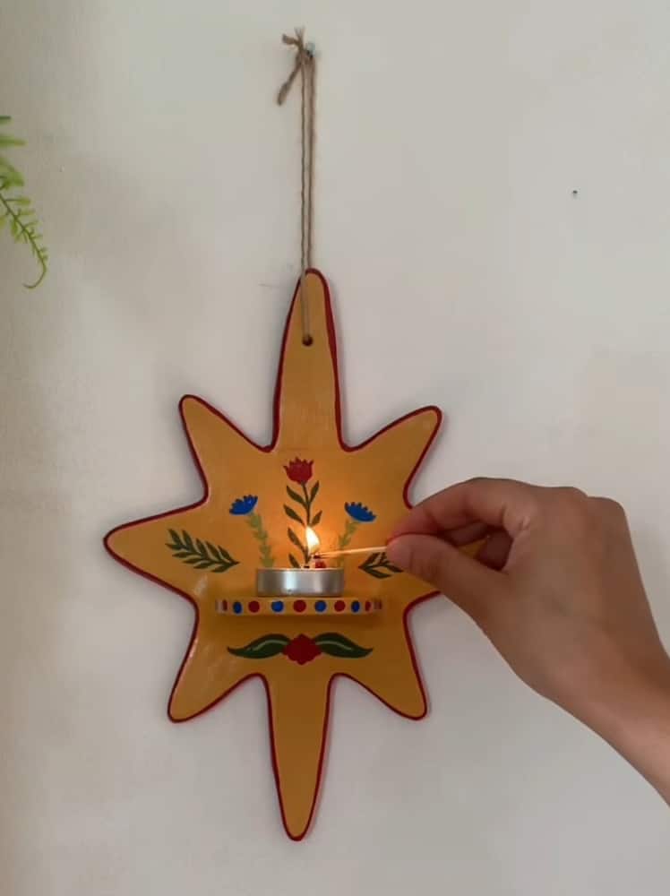 A hand lights a small candle placed on a yellow, star-shaped wall hanging with colorful floral designs. The decoration is attached to the wall with a brown string.