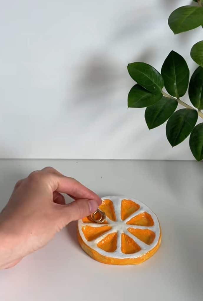 A hand places gold rings on a ceramic tray shaped and painted like an orange slice, with a green leafy plant in the background on a white surface.