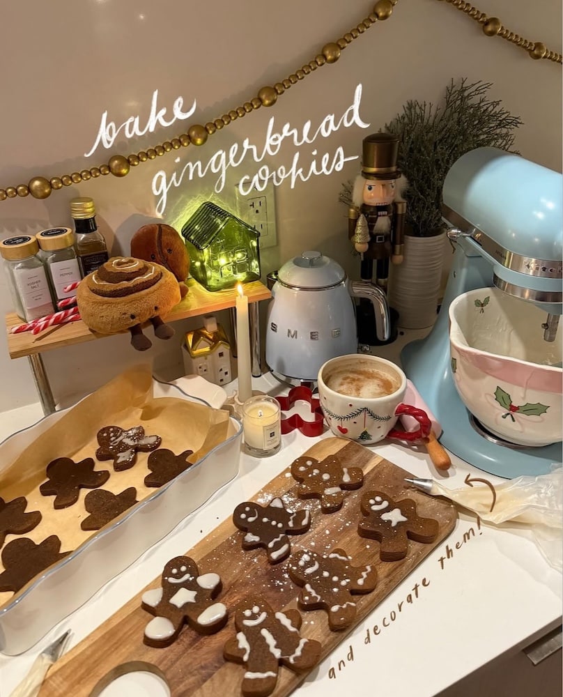 A cozy kitchen scene with gingerbread cookies—some decorated with icing—on a tray and counter. A blue mixer, mug of coffee, candles, and festive decor surround the baking area. Text reads bake gingerbread cookies and decorate them.