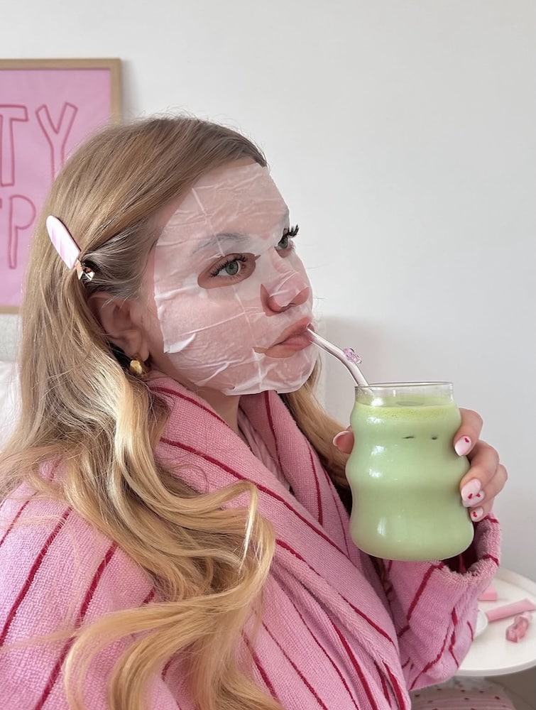 A woman wearing a sheet mask and pink robe sips a green drink through a straw. She has blonde hair with a pink hair clip and is sitting in a cozy setting with a pink-themed background.