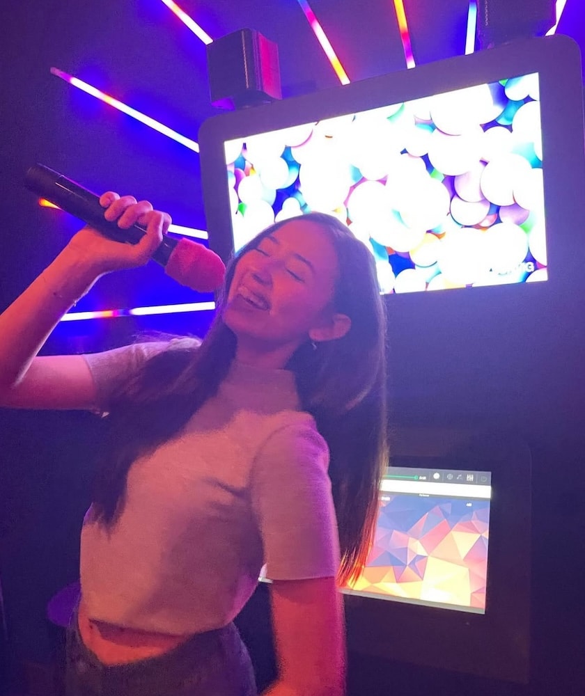 A woman with long hair joyfully sings into a microphone in front of a colorful, illuminated karaoke screen in a dark room. Neon lights create a vibrant atmosphere.