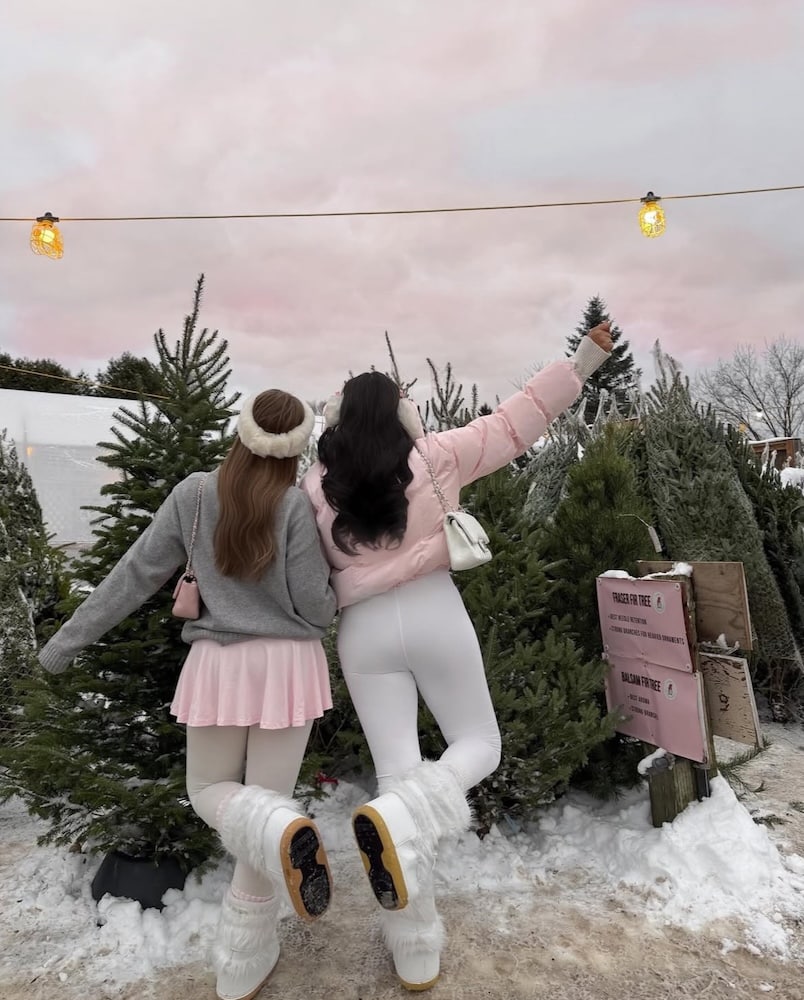 Two women in winter outfits pose playfully with their backs to the camera among snow-covered Christmas trees under a pink sky, with string lights glowing above them.