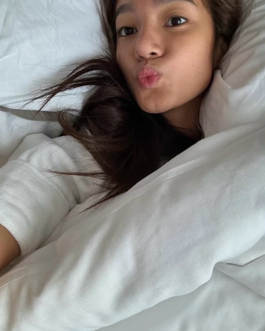 A young woman with long dark hair lies in bed under white blankets, resting her head on a pillow and making a playful kissy face at the camera—ready to share her favorite morning tips to help you become a morning person.