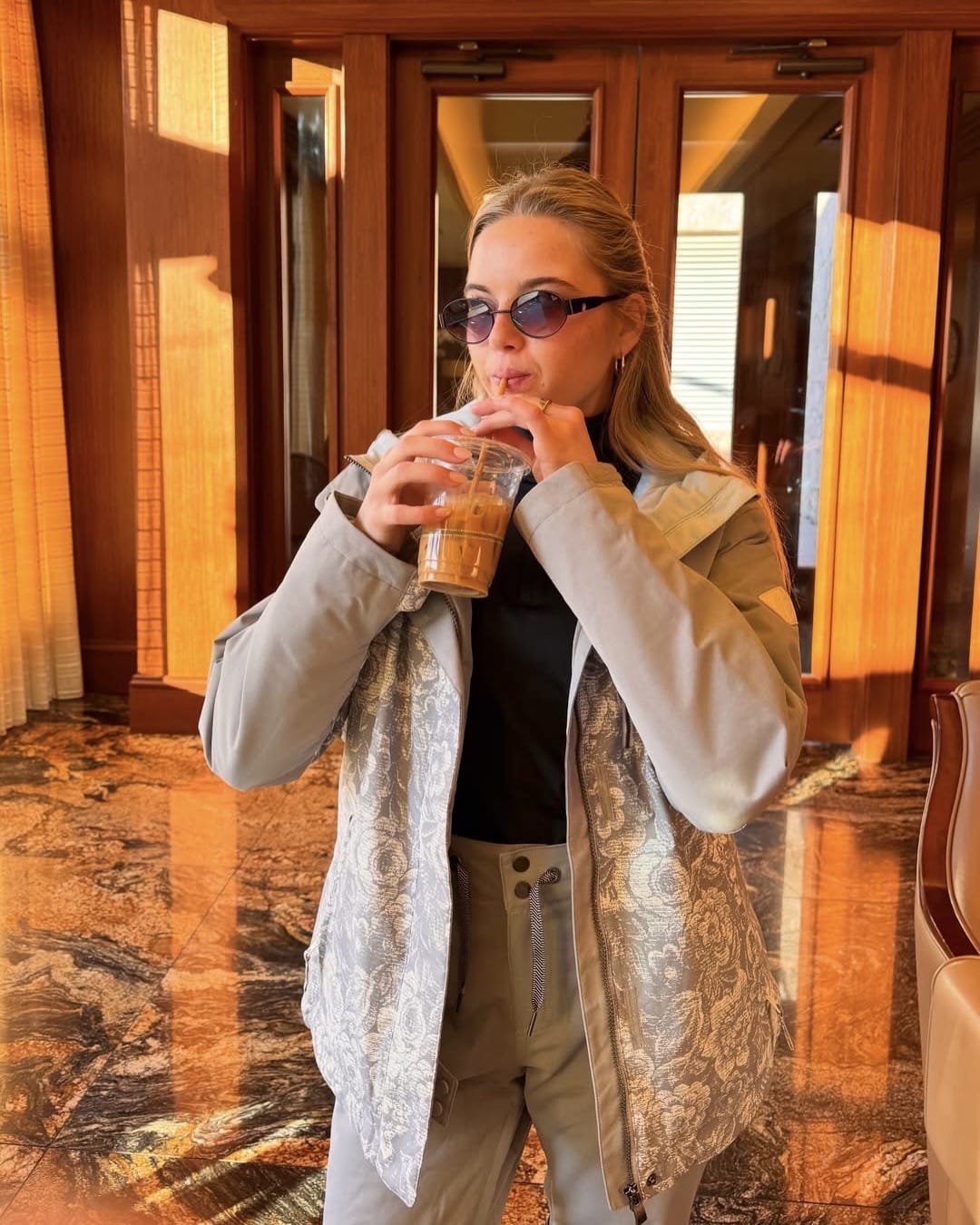 A morning person in chic sunglasses and a patterned jacket sips iced coffee through a straw while standing indoors in sunlight, with wooden doors and marble floors in the background.