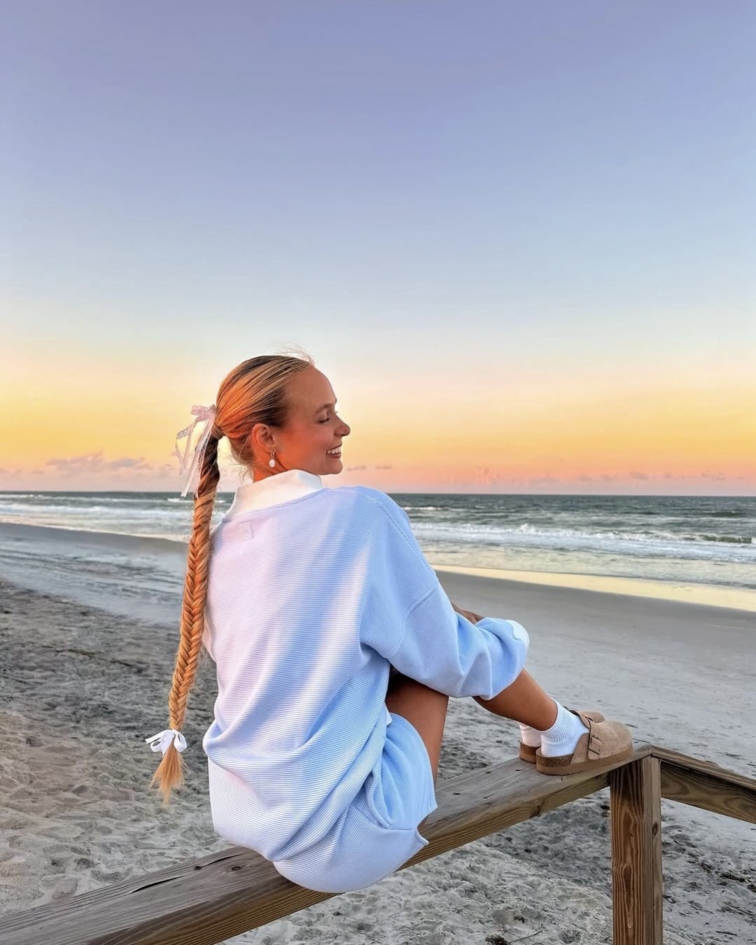 A woman with a long braided ponytail, wearing a light blue sweater and white socks, sits on a wooden railing by the beach at sunrise. Smiling as she looks at the ocean, she enjoys the calm—one of her favorite morning tips to become a morning person.