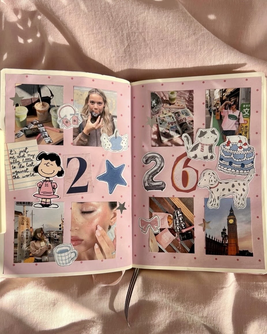 An open scrapbook with pink pages features collages of photos, cartoon stickers, doodles, and cut-out numbers “2 6.” Images include a woman, coffee, Big Ben, makeup, and playful decorations. Sunlight casts shadows on the book.