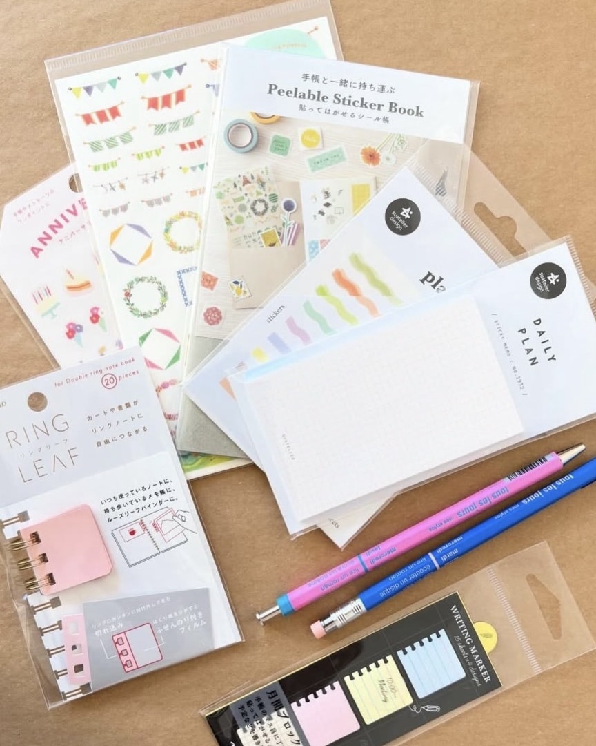 A variety of colorful stationery items, perfect for fueling your scrapbooking obsession, are arranged on a brown surface. Designs feature sticker sheets, sticky notes, pencils, and index tabs with banners, plants, and pastel colors.