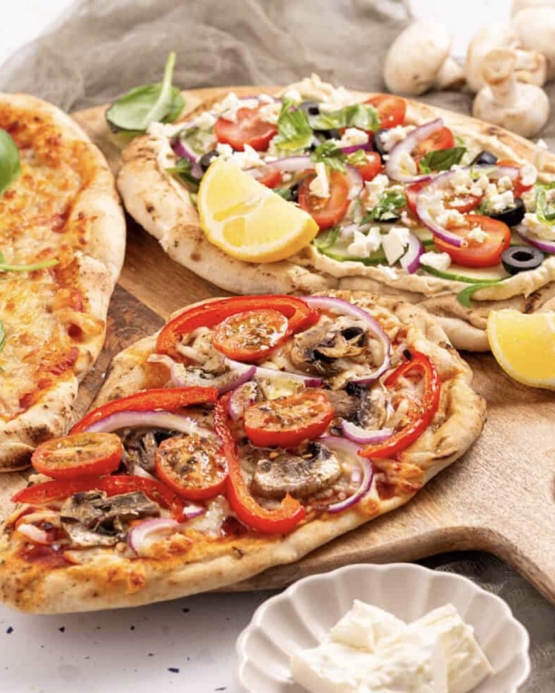Three flatbread pizzas on a wooden board: one topped with tomatoes, red onions, mushrooms, and red peppers; another with fresh veggies, olives, and feta; and a cheese pizza. Lemon wedges and feta cheese are nearby.